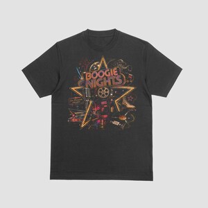 Op de afbeelding: Een antracietgrijs T-shirt met korte mouwen en een grote stervormige grafische print met de tekst "BOOGIE NIGHTS" in een retro lettertype. Het ontwerp bevat illustraties van een microfoon, filmrol, vinylplaat en een klassieke auto.