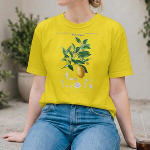 May include: A bright yellow t-shirt featuring a botanical illustration of a lemon branch with green leaves and a single yellow lemon. The graphic includes the text "Botanique" and smaller detailed diagrams of plant parts printed in blue ink.