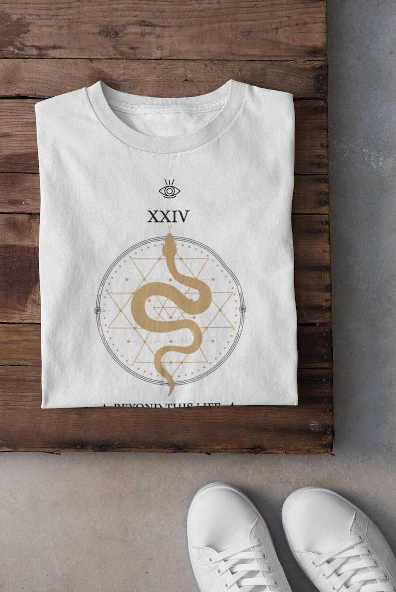 May include: White t-shirt with a gold snake design inside a geometric shape. The shirt has the number XXIV and an eye symbol above the snake. The words "A BEYOND THIS LIFE" are printed below the design. The shirt is on a wooden surface.