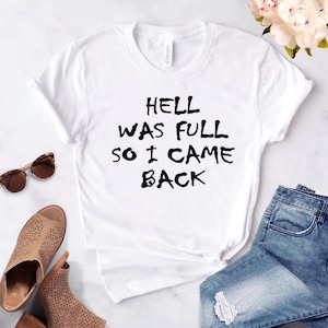 Może przedstawiać: Biała koszulka z czarnym napisem "Piekło było pełne, więc wróciłem".