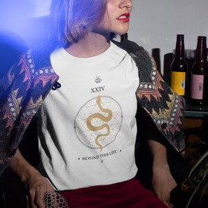 May include: White t-shirt featuring a gold snake design within a geometric pattern. The shirt includes the text "XXIV" above the design and "BEYOND THIS LIFE" below. The shirt is paired with a red skirt and a patterned jacket.