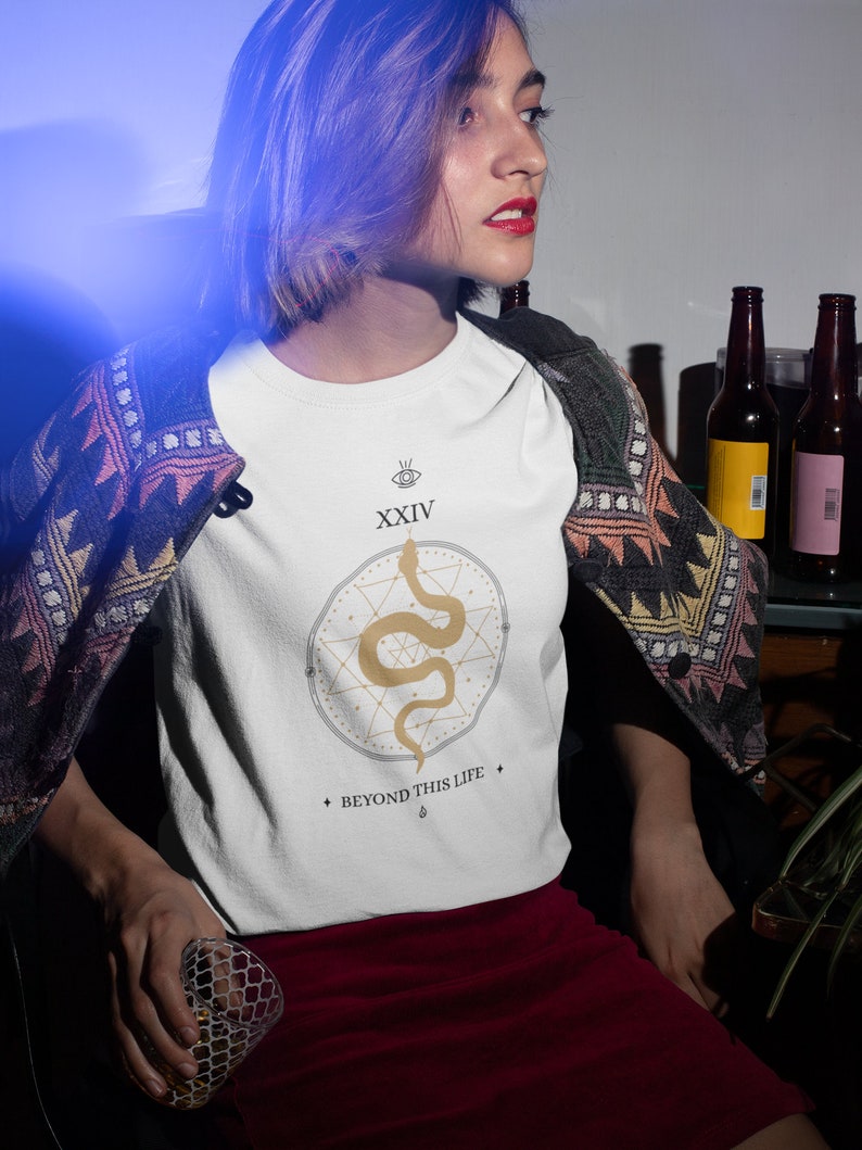 May include: White t-shirt featuring a gold snake design within a geometric pattern. The shirt includes the text "XXIV" above the design and "BEYOND THIS LIFE" below. The shirt is paired with a red skirt and a patterned jacket.