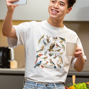 May include: A white t-shirt with a colorful print of various birds. The person wearing the shirt is smiling and holding a white mug in one hand and a smartphone in the other.