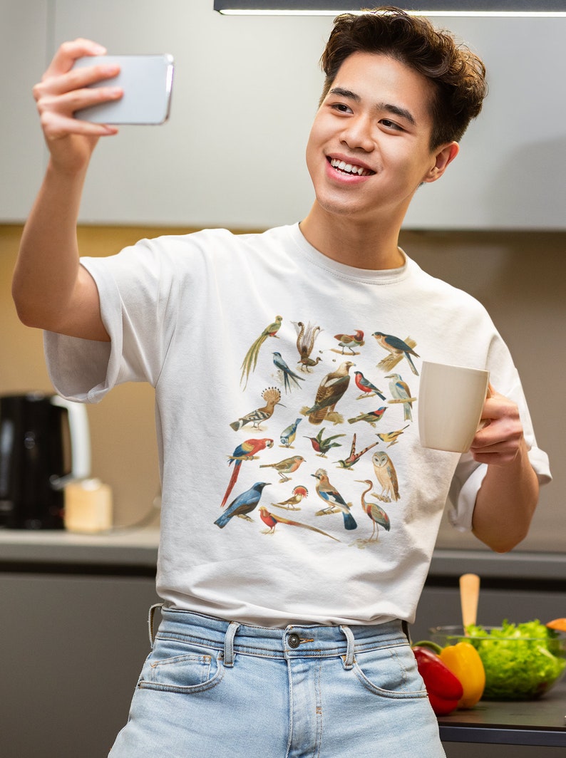 May include: A white t-shirt with a colorful print of various birds. The person wearing the shirt is smiling and holding a white mug in one hand and a smartphone in the other.