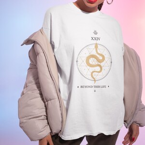 May include: White t-shirt with a gold snake design inside a circle, the number XXIV, and the text "BEYOND THIS LIFE". The shirt is worn with a beige puffer jacket and dark brown pants.