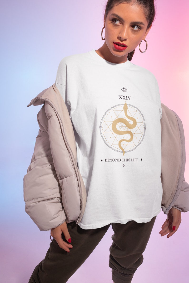 May include: White t-shirt with a gold snake design inside a circle, the number XXIV, and the text "BEYOND THIS LIFE". The shirt is worn with a beige puffer jacket and dark brown pants.