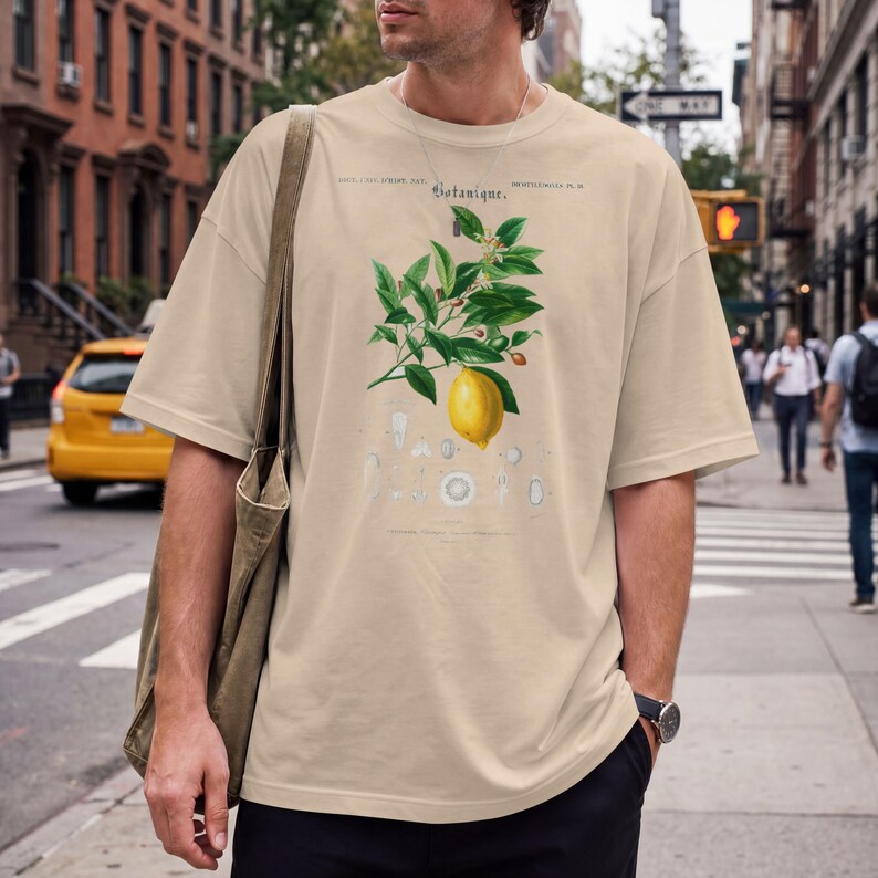 May include: A beige short-sleeved t-shirt featuring a botanical illustration of a lemon branch with green leaves and a yellow lemon. The text "Botanique" is printed above the illustration, accompanied by smaller, faint botanical diagrams below.
