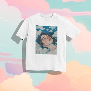 May include: White t-shirt with a graphic print of a woman with long brown hair lying in clouds. The woman is looking up at the sky with her eyes closed.