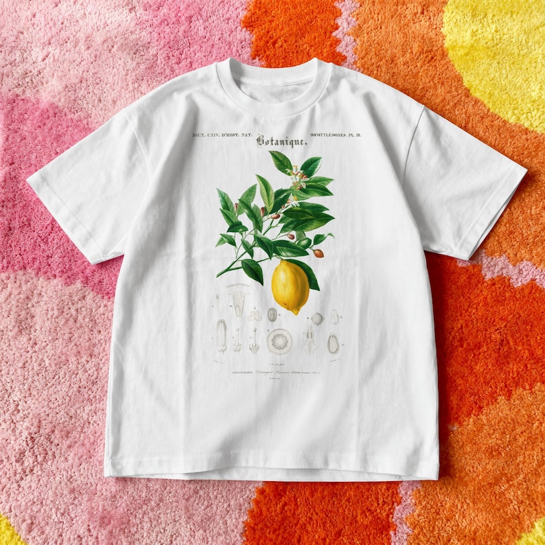 May include: A white short-sleeve t-shirt featuring a botanical illustration of a lemon branch with green leaves and a single yellow lemon. The graphic includes the text "Botanique" and smaller scientific diagrams of plant parts below the fruit.