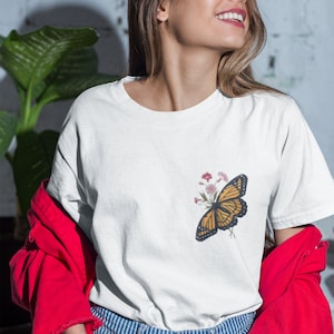 Puede incluir: Una camiseta blanca con un dise&ntilde;o de mariposa y flores. La mariposa es amarilla y negra con vetas marrones. Las flores son rosas y blancas.