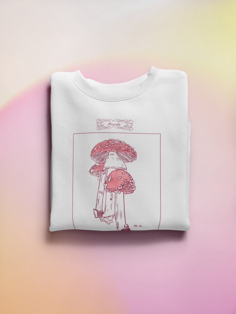 K&ouml;nnte beinhalten: Wei&szlig;es Sweatshirt mit einer Illustration von rosa und roten Pilzen und dem Text "Amanda" in einem dekorativen Rahmen.