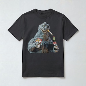 May include: A black short-sleeved t-shirt featuring a graphic print of a grey, textured monster with sharp teeth. The monster holds a red pack of cigarettes and a glass bottle of Coca-Cola, with a lit cigarette hanging from its mouth.