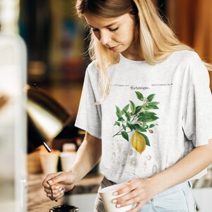 May include: A light gray short-sleeve t-shirt featuring a botanical illustration of a yellow lemon on a branch with green leaves and small white flowers. The text Botanique is printed above the lemon graphic in a dark, elegant serif font.