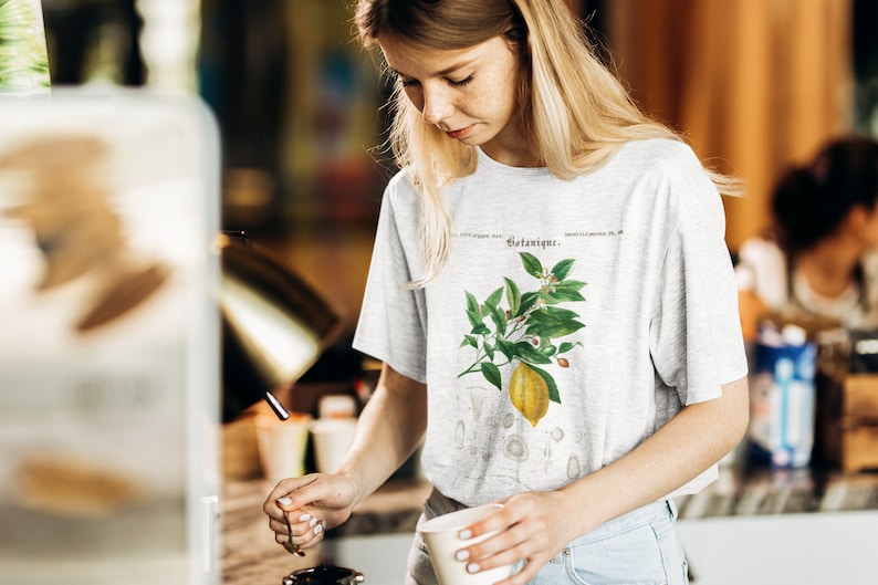 May include: A light gray short-sleeve t-shirt featuring a botanical illustration of a yellow lemon on a branch with green leaves and small white flowers. The text Botanique is printed above the lemon graphic in a dark, elegant serif font.