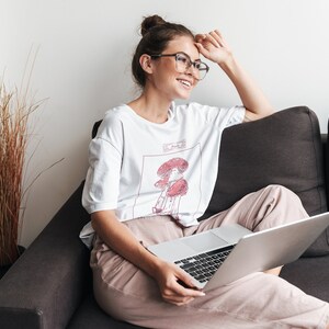 K&ouml;nnte beinhalten: Eine Frau mit Brille sitzt auf einem Sofa mit einem Laptop auf dem Scho&szlig;. Sie tr&auml;gt ein wei&szlig;es T-Shirt mit einem Pilzmotiv und dem Text "Shroom Queens".