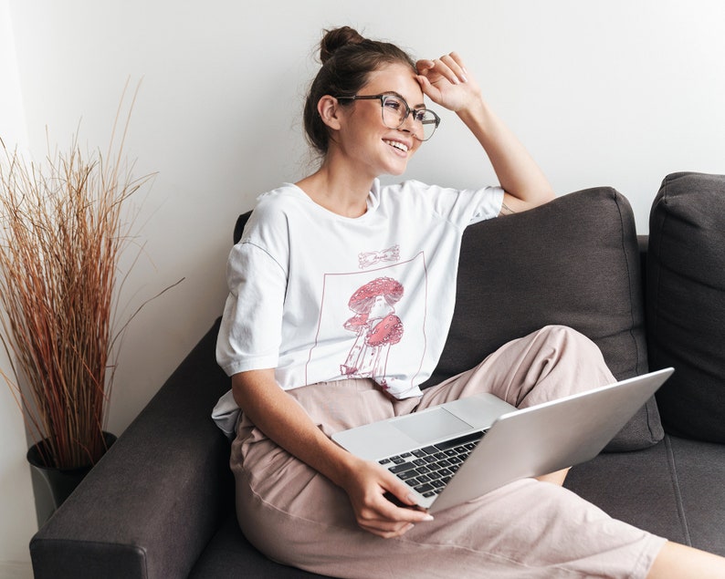 K&ouml;nnte beinhalten: Eine Frau mit Brille sitzt auf einem Sofa mit einem Laptop auf dem Scho&szlig;. Sie tr&auml;gt ein wei&szlig;es T-Shirt mit einem Pilzmotiv und dem Text "Shroom Queens".