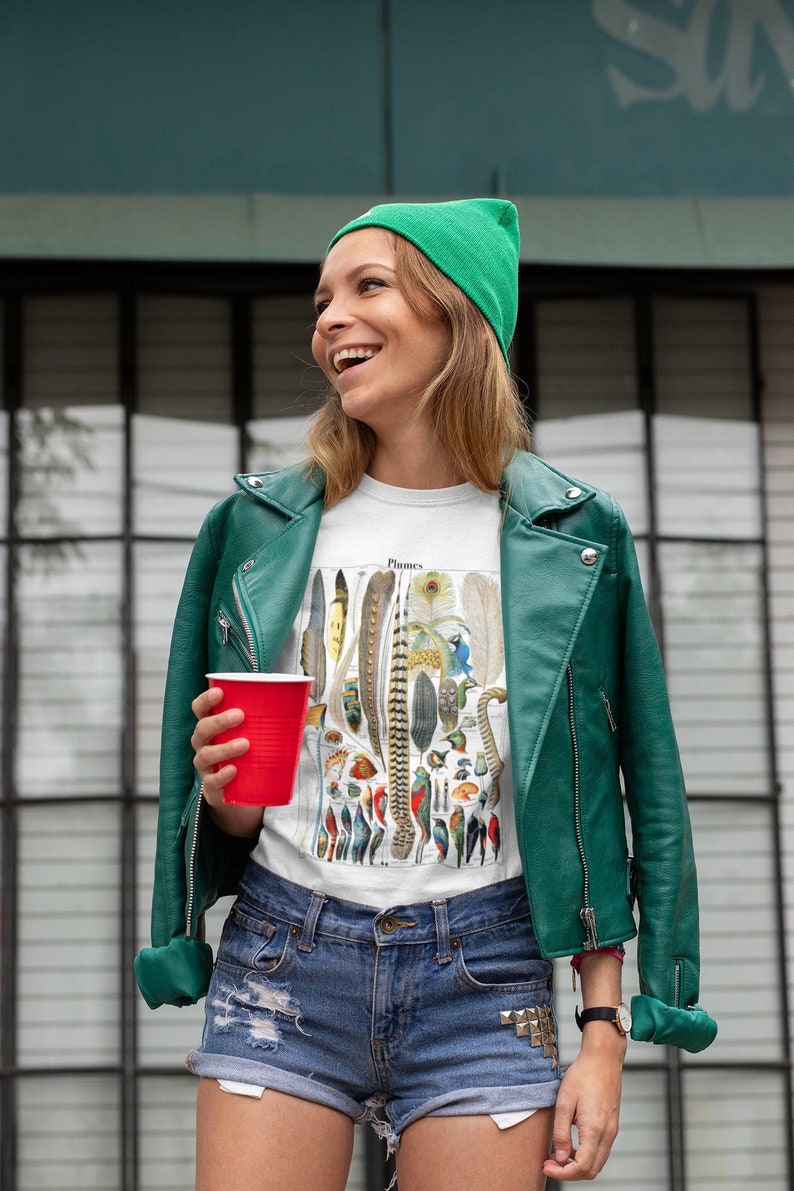 May include: A woman wearing a green beanie, a white t-shirt with a vintage illustration of feathers and birds, a green leather jacket, and denim shorts. The t-shirt has the word "Plumes" printed on it.