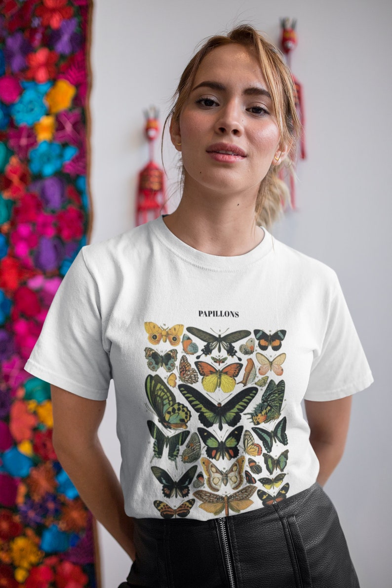 Może przedstawiać: Biały t-shirt z vintage'ową ilustracją motyli. Ilustracja jest w odcieniach brązu, zieleni, ż&oacute;łci i błękitu. Tekst "PAPILLONS" jest nadrukowany nad ilustracją.