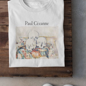 May include: White t-shirt featuring a print of a painting by Paul Cézanne depicting three skulls on a patterned fabric.