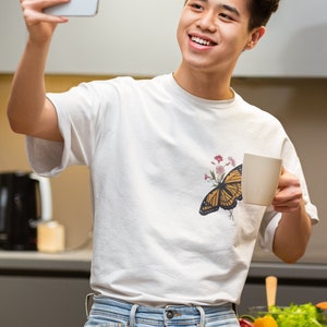 Puede incluir: Un joven con una camiseta blanca con estampado de mariposa y flores, pantalones vaqueros azules, sosteniendo una taza blanca en una mano y un tel&eacute;fono inteligente en la otra. Est&aacute; sonriendo y mirando el tel&eacute;fono inteligente.