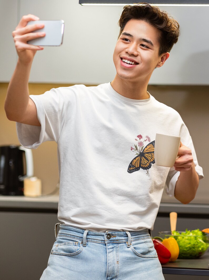 Puede incluir: Un joven con una camiseta blanca con estampado de mariposa y flores, pantalones vaqueros azules, sosteniendo una taza blanca en una mano y un tel&eacute;fono inteligente en la otra. Est&aacute; sonriendo y mirando el tel&eacute;fono inteligente.