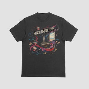 Op de afbeelding: Een antracietgrijs T-shirt met korte mouwen en een grafische print met de tekst PUNCH-DRUNK LOVE. Het ontwerp bevat een hoofdloos figuur in een pak, een harmonium, rode bekers, verspreide kaartjes en een open deur naar een strand.