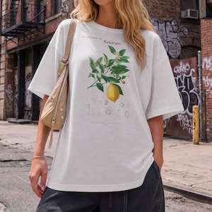 May include: A white short-sleeved t-shirt featuring a botanical illustration of a lemon branch with green leaves and a single yellow lemon. The graphic includes the text "Botanique" and smaller scientific diagrams printed in black ink.