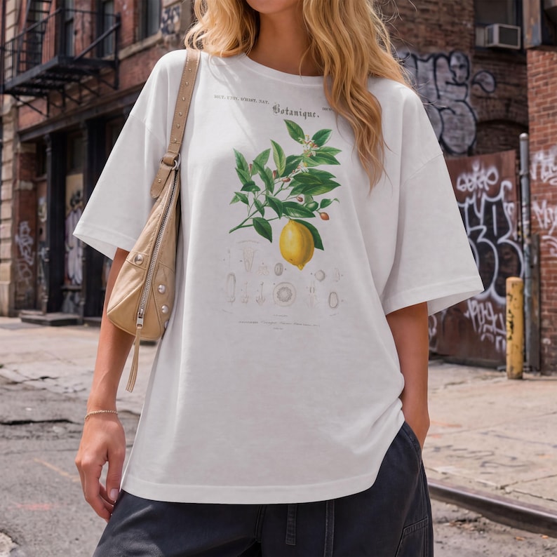 May include: A white short-sleeved t-shirt featuring a botanical illustration of a lemon branch with green leaves and a single yellow lemon. The graphic includes the text "Botanique" and smaller scientific diagrams printed in black ink.