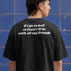 Op de afbeelding: Een zwart t-shirt met korte mouwen en witte tekst op de achterkant die luidt: if i go to hell at least i'll be with all my friends. Het kledingstuk heeft een ronde hals en een relaxte pasvorm, waardoor de belettering duidelijk zichtbaar is.