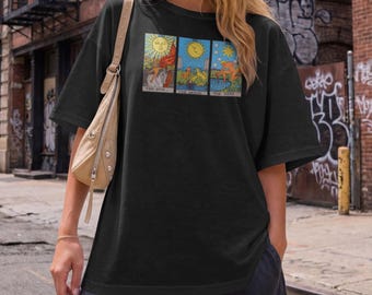 May include: A black short-sleeved t-shirt featuring a graphic print of three tarot cards labelled The Sun, The Moon, and The Star. The design displays celestial imagery with a sun, moon, and star, alongside figures in a colourful, detailed illustration.