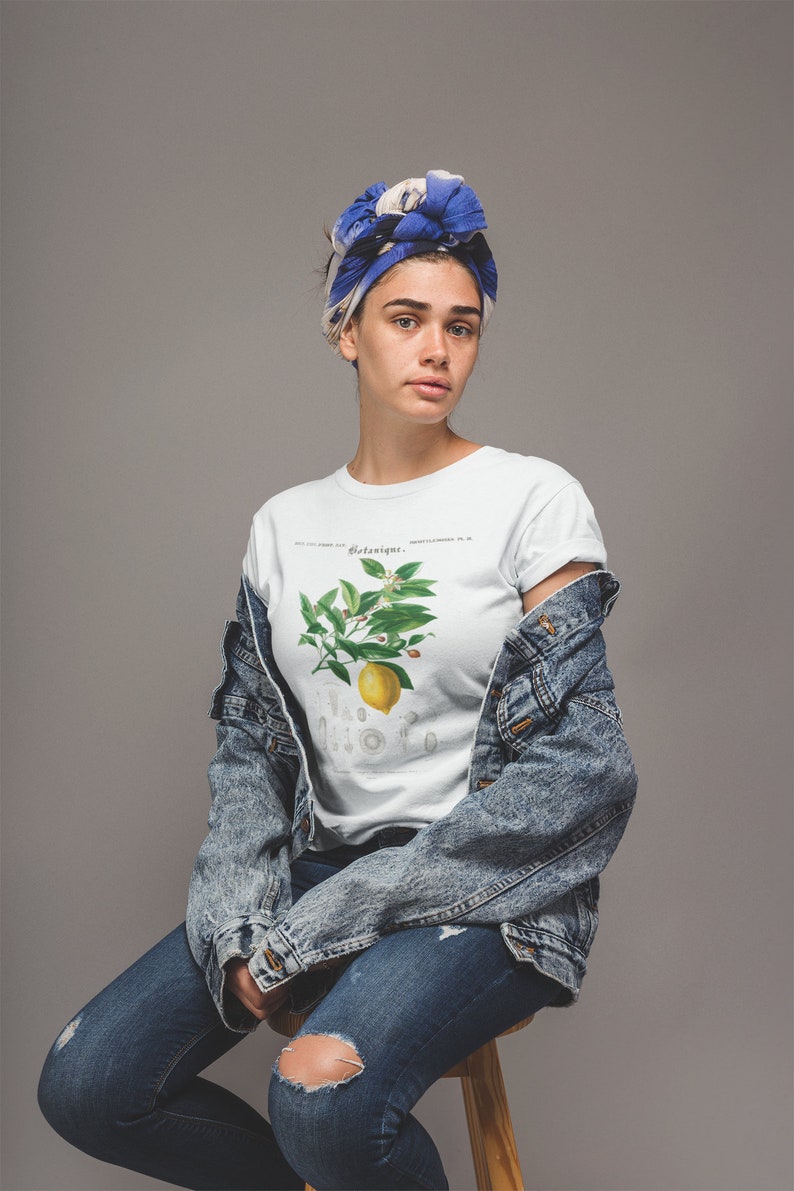 May include: A white crew neck t-shirt featuring a botanical illustration of a lemon branch with green leaves and a yellow lemon, with the text Botanique printed above the graphic, worn with a blue acid-wash denim jacket and blue jeans.