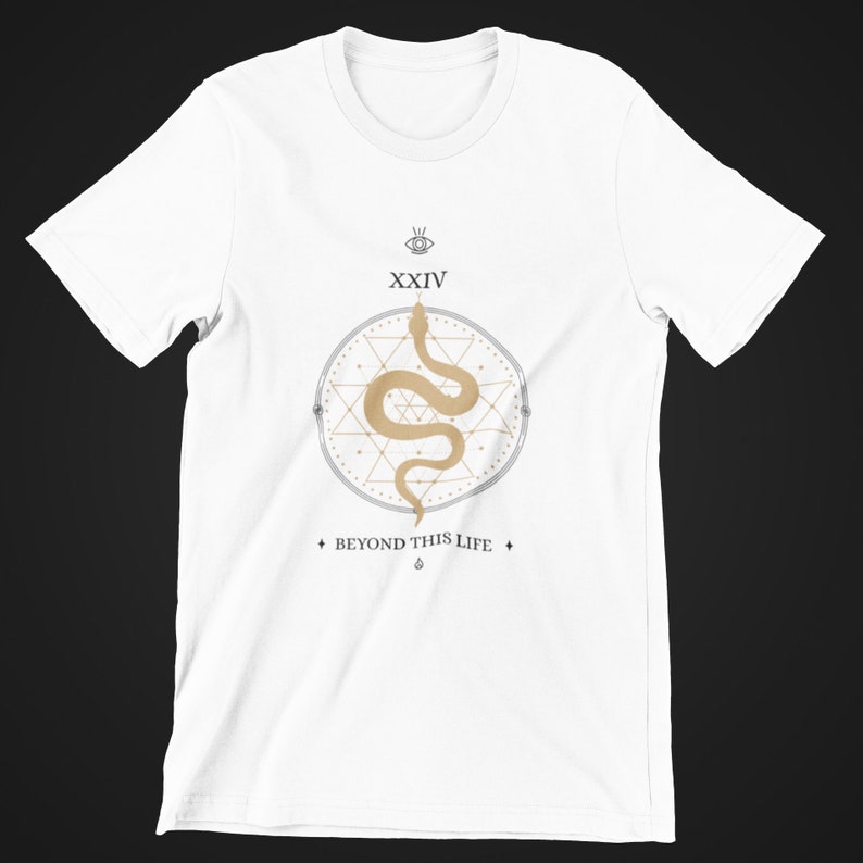 May include: White t-shirt featuring a circular design with a beige snake, geometric shapes, and the text "XXIV" and "BEYOND THIS LIFE". The design includes an eye symbol and a diamond shape. The shirt is made of a soft material.