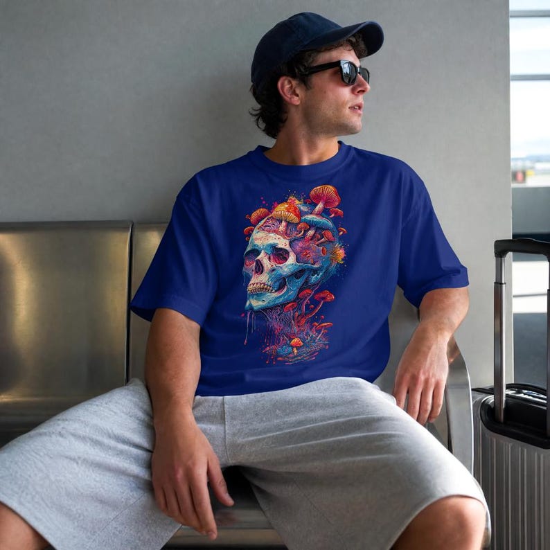 Op de afbeelding: Een koningsblauw t-shirt met korte mouwen en een levendige grafische print van een menselijke schedel met kleurrijke paddenstoelen die uit de bovenkant en de basis groeien. Het ontwerp bevat tinten rood, oranje, geel en blauw.