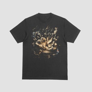 Op de afbeelding: Een antracietgrijs t-shirt met korte mouwen en een centrale grafische print van een grote witte magnolia-bloem, omringd door donkere, atmosferische elementen en het woord MAGNOLIA in een schreeflettertype boven de bloem.