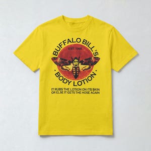 May include: A bright yellow short-sleeve t-shirt featuring a graphic of a moth with a skull pattern on its back, centred within a red circle. The text reads BUFFALO BILL'S BODY LOTION EST. 1991 and IT RUBS THE LOTION ON ITS SKIN OR ELSE IT GETS THE HOSE AGAIN.