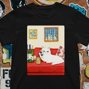以下が含まれることがあります： 赤いソファに座り、ワイングラスを持った白い猫のイラストが描かれた黒いTシャツ。ワインボトル、額入りの写真、瓶に書かれた「The Red Book」の文字が含まれています。テーマはワインと猫です。