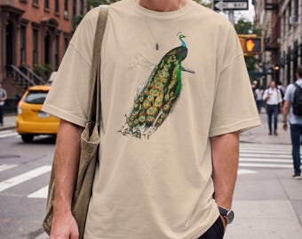 May include: A beige short-sleeve t-shirt featuring a detailed graphic illustration of a peacock with vibrant green, blue, and gold plumage perched on a branch. The shirt has a crew neckline and a relaxed fit, paired with a dark olive green tote bag.