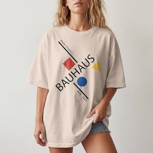 May include: An oversized beige t-shirt featuring a graphic design with the text Walter Gropius, BAUHAUS, and 1919-1923. The print includes a red square, a blue circle, and a yellow triangle arranged along black diagonal lines.