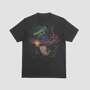 Op de afbeelding: Een antracietgrijs T-shirt met korte mouwen en een grafische print van een strand bij zonsondergang, een notitieblok en een goudkleurig object. De tekst "INHERENT VICE" staat in neongroen en "Surfside MOTEL" in roze neonletters.
