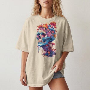 Op de afbeelding: Een beige t-shirt met korte mouwen en een grote, kleurrijke grafische print op de voorkant. Het ontwerp toont een blauwe menselijke schedel met diverse rode, oranje en roze paddenstoelen die uit de bovenkant en de basis groeien.
