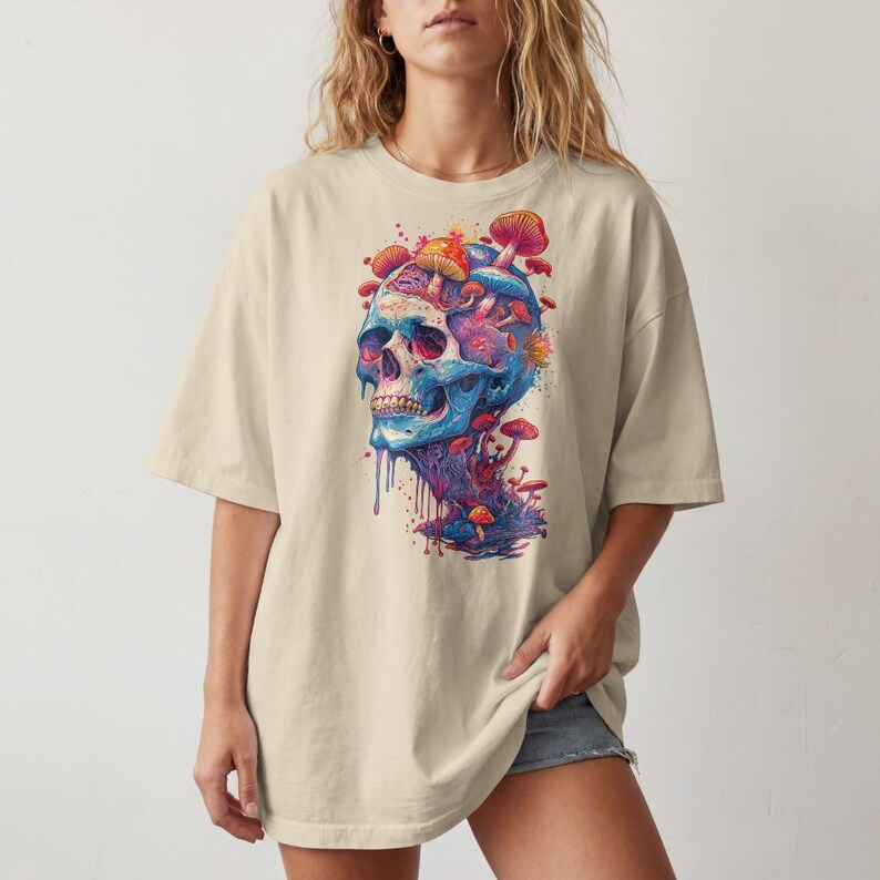 Op de afbeelding: Een beige t-shirt met korte mouwen en een grote, kleurrijke grafische print op de voorkant. Het ontwerp toont een blauwe menselijke schedel met diverse rode, oranje en roze paddenstoelen die uit de bovenkant en de basis groeien.