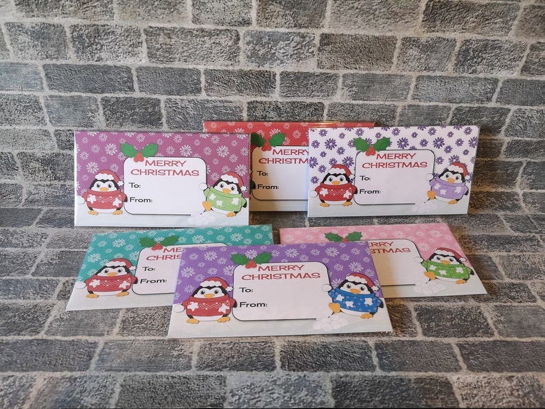 May include: Set of six purple, pink, blue, and green gift tags with a penguin wearing a red, green, or purple sweater. Each tag has a snowflake and flower pattern and says "Merry Christmas" with a space for "To:" and "From:".