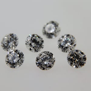 May include: Eight clear round faceted gemstones, likely diamonds, on a white surface.