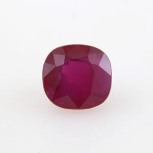 May include: A cushion-cut ruby gemstone with a deep red hue. The gemstone is faceted, displaying multiple planes that reflect light. The ruby is set against a white background, highlighting its color and clarity.