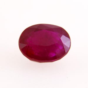 May include: A faceted, oval-cut ruby gemstone. The gem is a rich, deep red hue, with a polished surface. The ruby is displayed against a white backdrop, accentuating its colour and clarity.