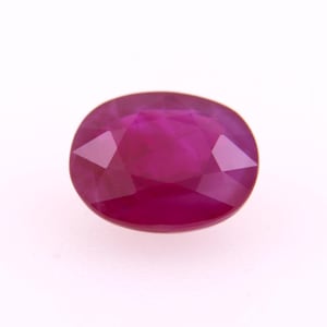 May include: A faceted, oval-shaped ruby gemstone. The gem is a deep, saturated red color with a polished surface. The facets create a play of light and shadow, enhancing the stone's brilliance. The ruby is set against a white background.