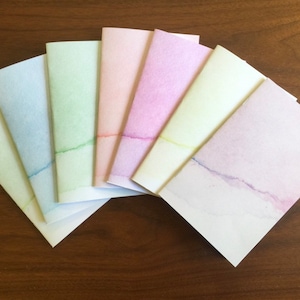 May include: Seven small, blank notebooks with watercolor painted edges in shades of blue, green, pink, and purple. The notebooks are arranged in a fan shape.