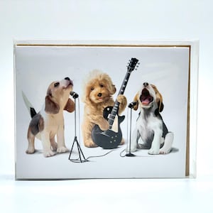 May include: Greeting card featuring three dogs as a rock band. A poodle plays a black electric guitar, while two beagles sing into microphones. The card is encased in a clear plastic sleeve, with a white background.