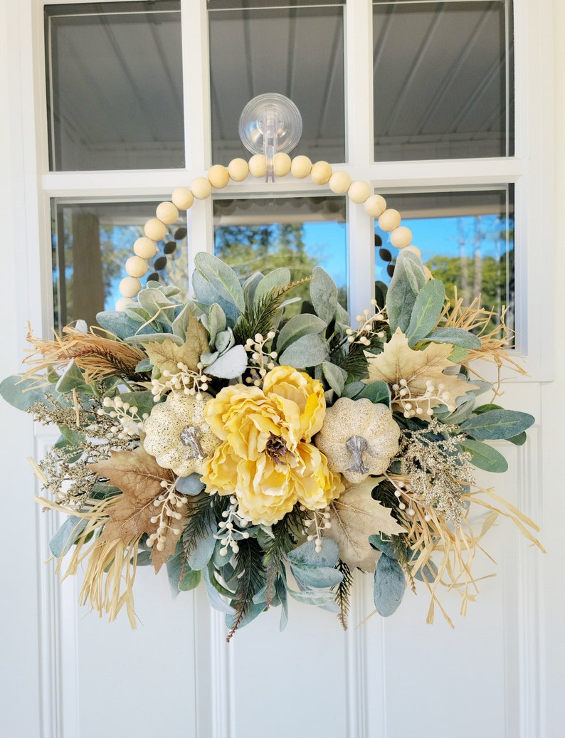 May include: A fall-themed wreath with a wooden bead base. The wreath is decorated with yellow flowers, white pumpkins, and green and brown foliage.