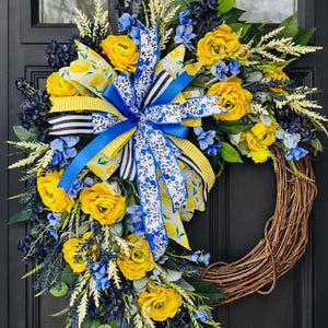 May include: A decorative wreath featuring yellow roses, blue flowers, and greenery. The wreath has a large bow with blue, yellow, and black and white striped ribbons. The wreath is made of woven brown branches.